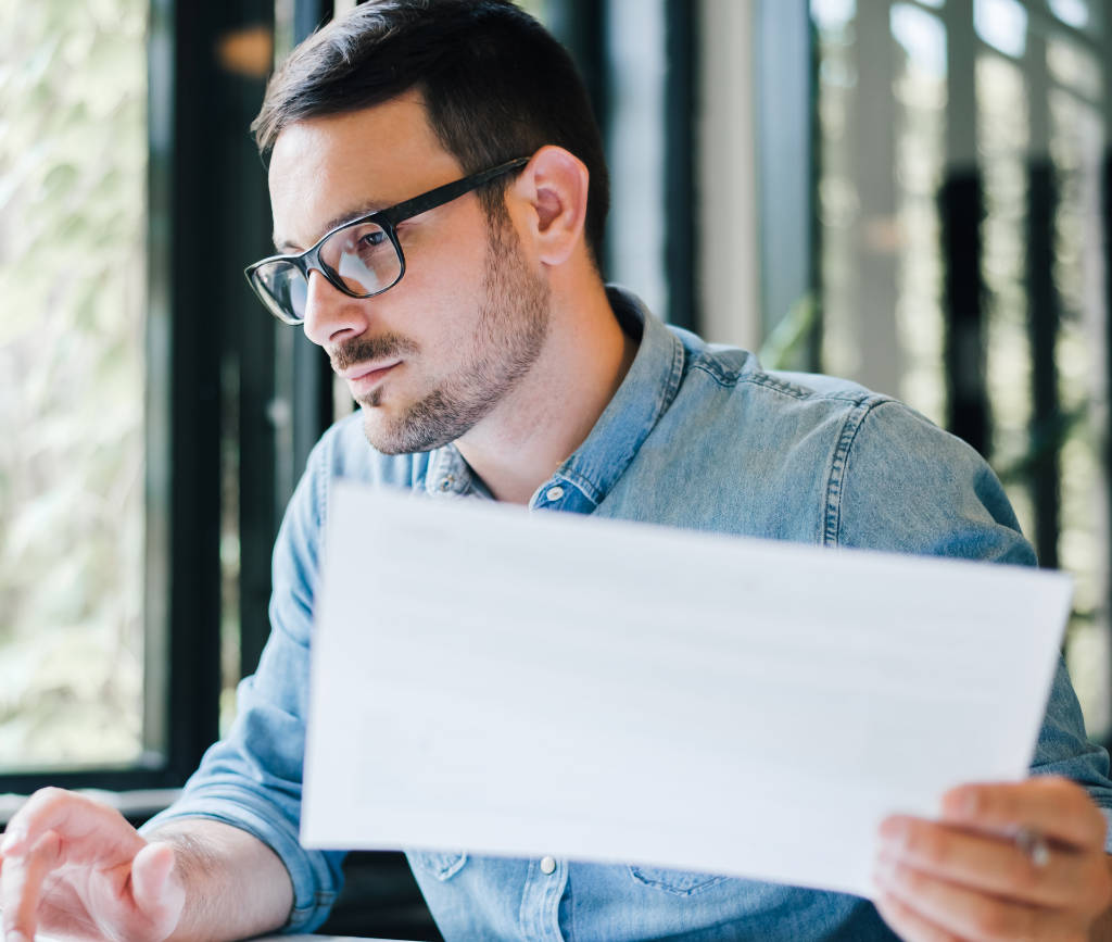 Man with glasses holding a paper in one hand and tapping a mousepad with other hand