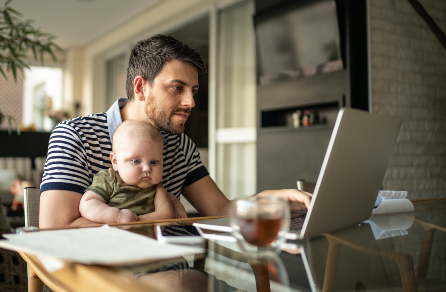 Man holding baby on lap, sitting at a glass table with paper on it and looking at laptop