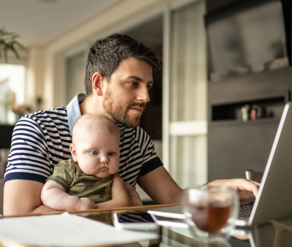 Man holding baby on lap, sitting at a glass table with paper on it and looking at laptop